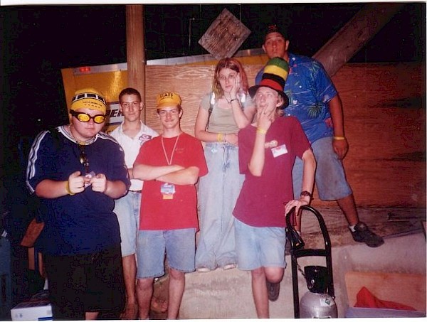 Group photo. Left to right: Jeeves in a yellow cap and yellow sunglasses pointing at the camera like the cool '90s dude he was; Matt in a white polo; Luke in a yellow cap and red shirt; Katt with pinkish hair, green top, jeans, leaning on my shoulder; me in a rainbow-colored tall hat, maroon shirt, hand on my chin looking thoughtfully upwards; and a dude I do not remember in a blue shirt