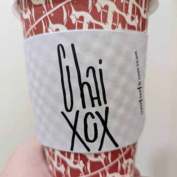 A white coffee cup sleeve that the barista has written 'Chai XCX' in black marker